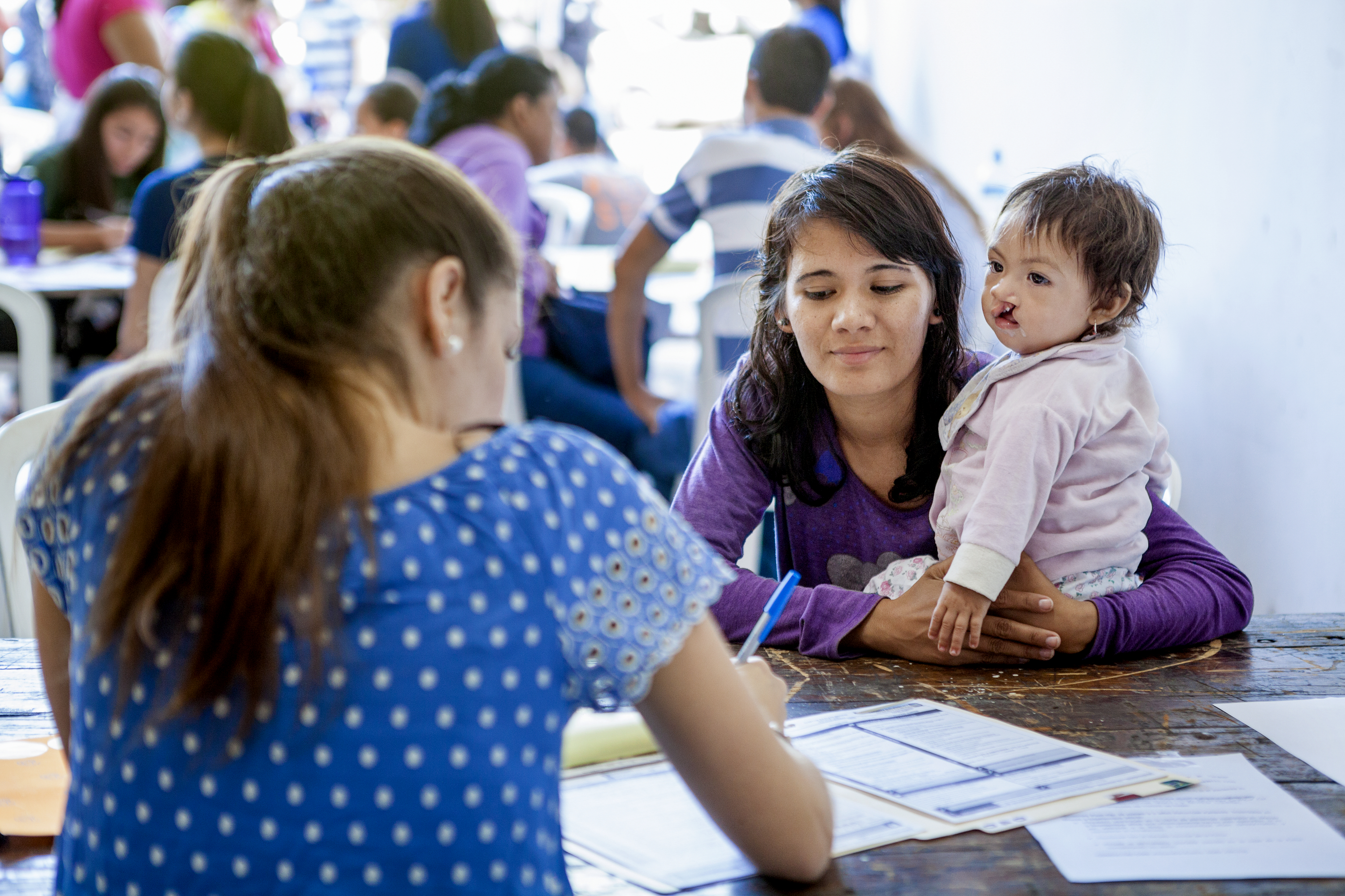 En kvinna med ett litet barn sitter mittemot en annan kvinna som fyller i papper vid ett bord med blanketter inomhus.