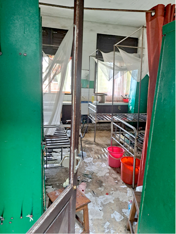 Ett rum med två metallängar, myggnät, papper på golvet, röda hinkar och en grön avskärmning, likt ett sjukhus eller studentrum.