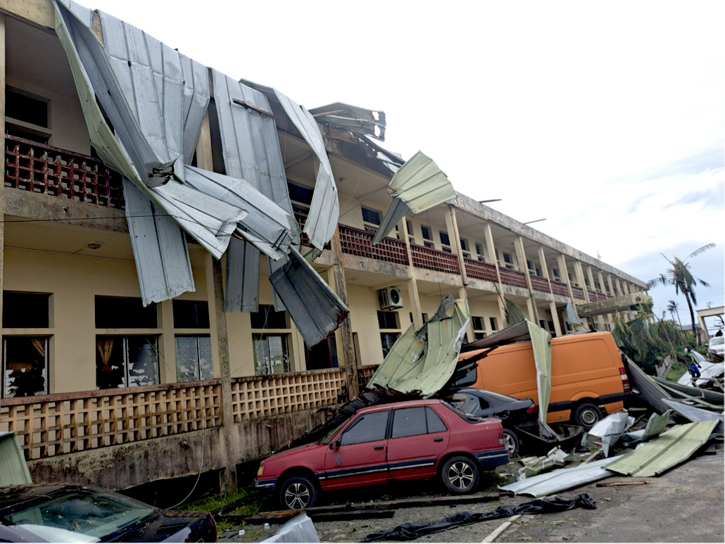 En byggnad med delvis borttaget tak, metallrester ligger på parkerade bilar och marken, vilket tyder på storm- eller vindskada.
