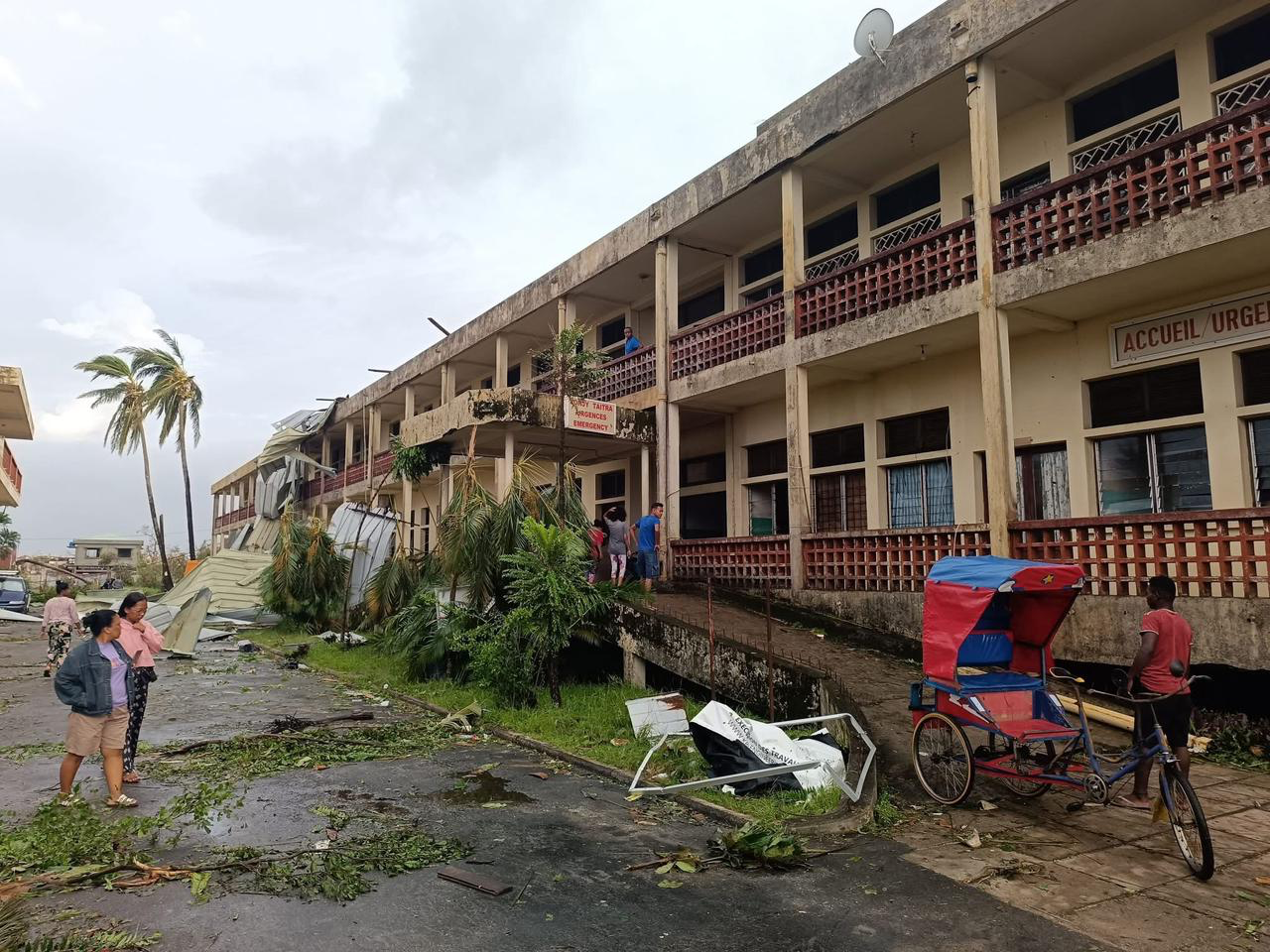 Personer står utanför en tvåvåningsbyggnad med skräp på marken. En trehjuling och böjda palmer syns i bilden.