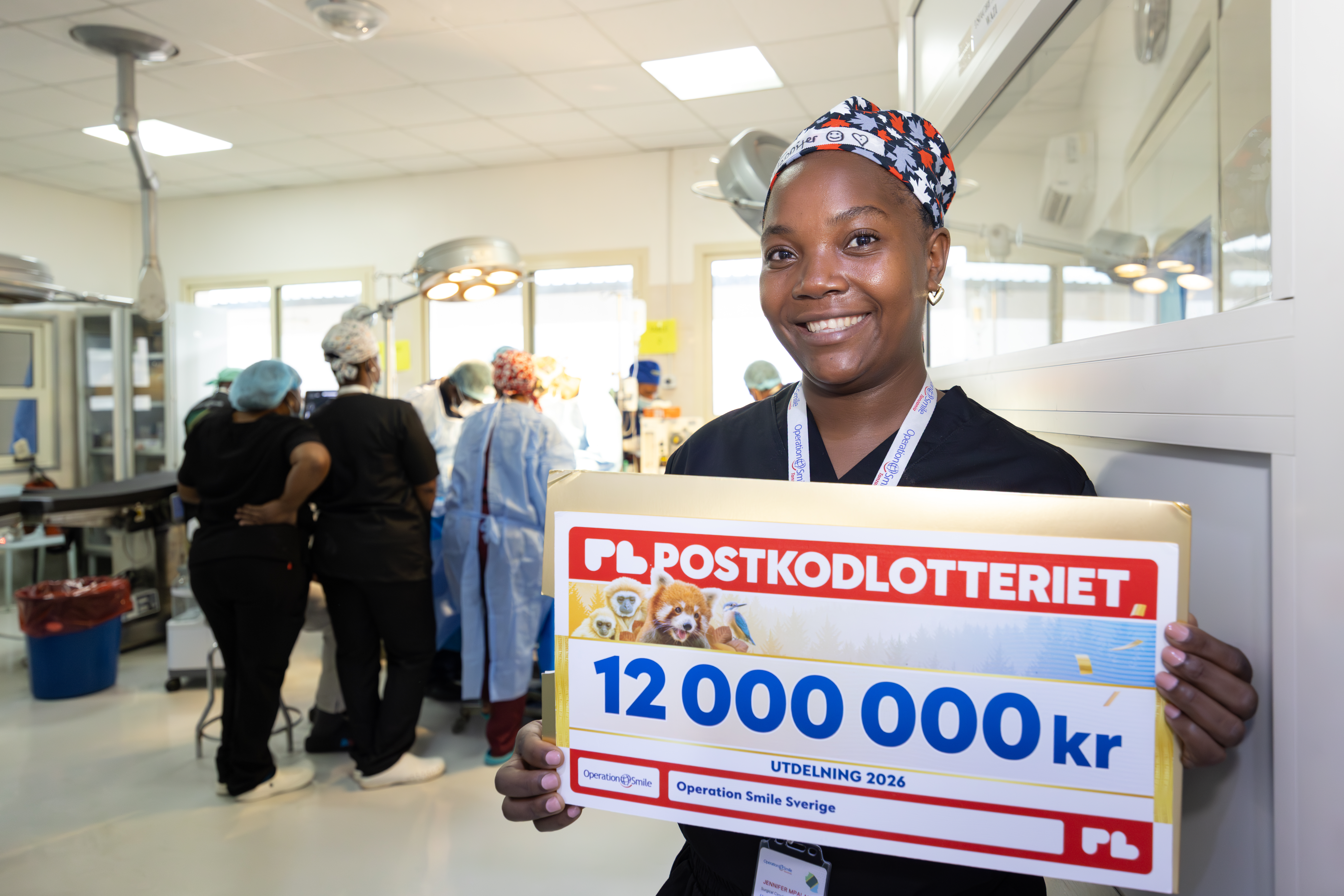 Kvinna i operationskläder håller en stor check på 12 000 000 kr från Postkodlotteriet medan annan personal arbetar i bakgrunden. Hon står utanför en operationssal.