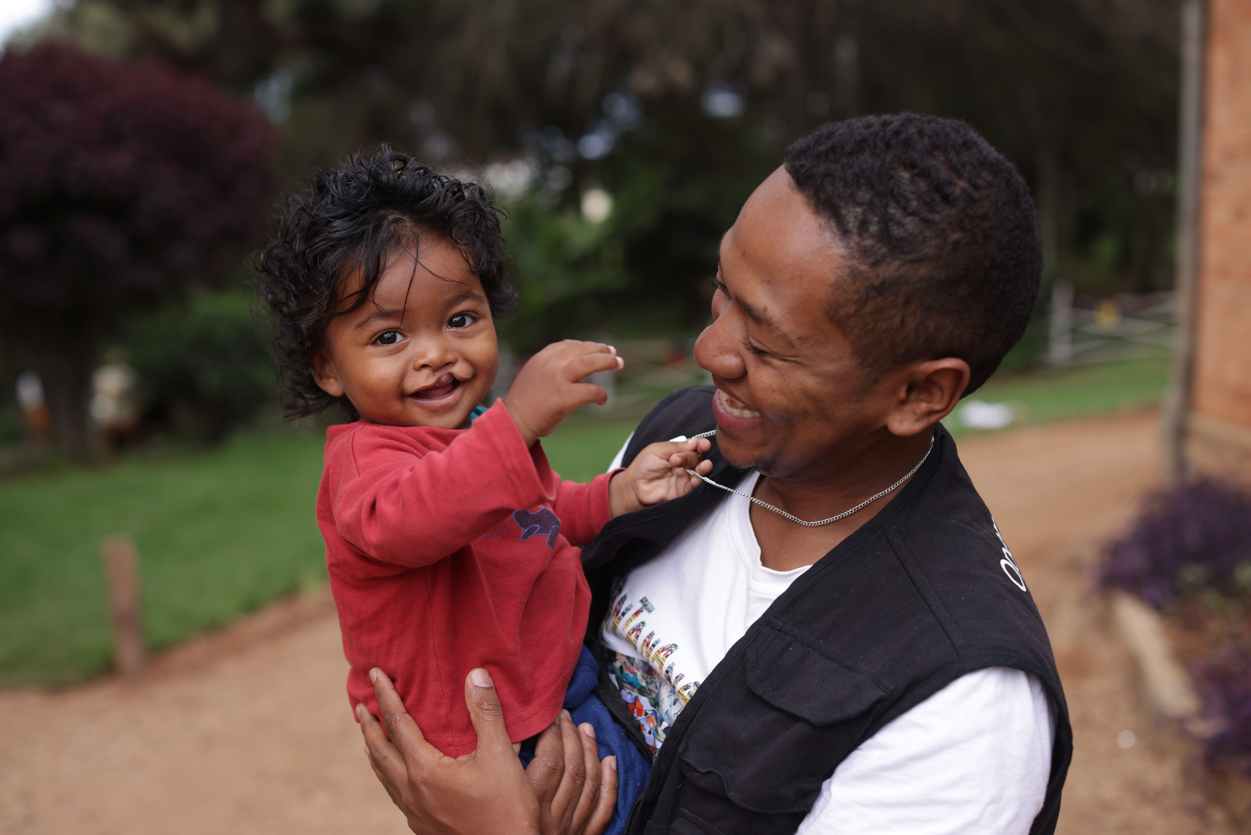 En man håller ett litet barn i famnen utomhus. Barnet sträcker sig mot mannens halskedja.