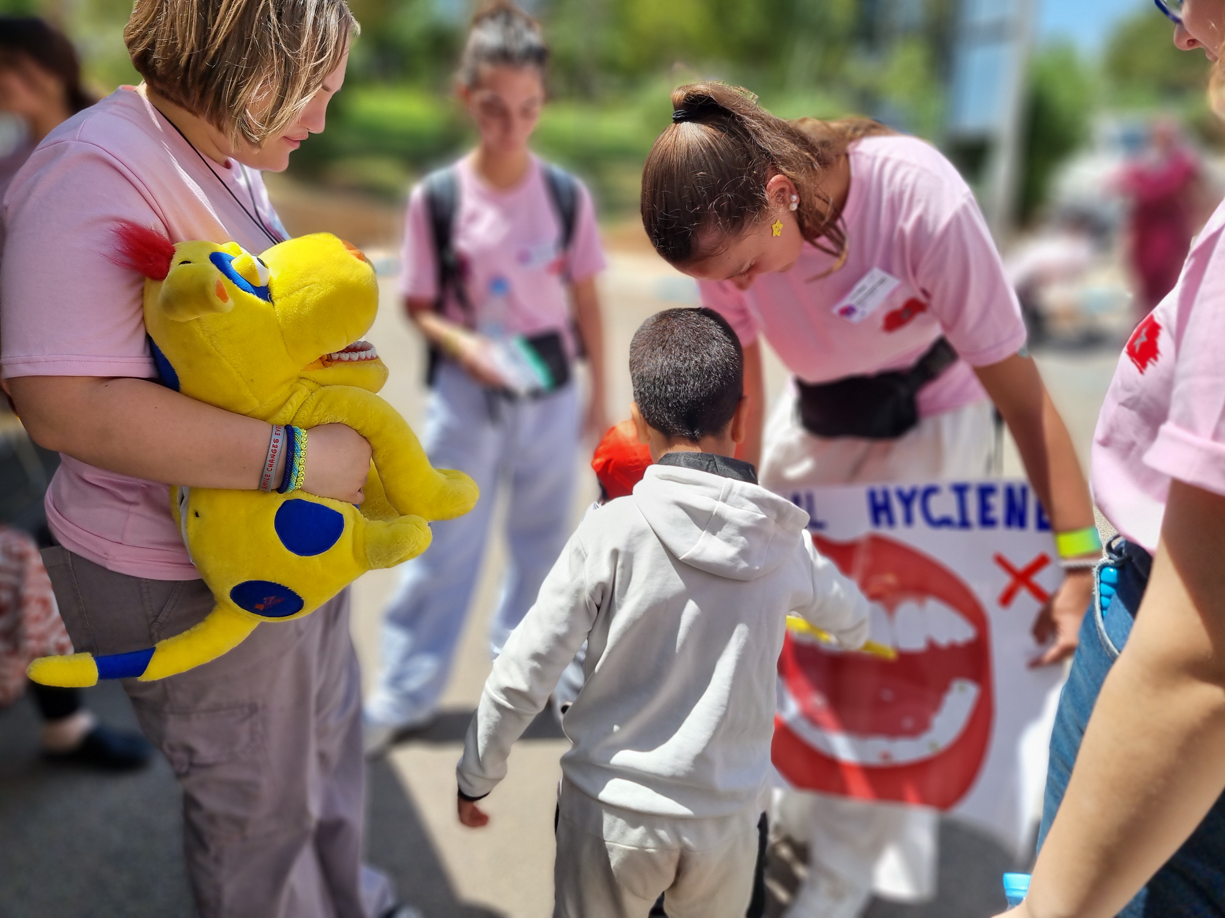 En grupp personer i rosa tröjor interagerar med ett barn. En håller i ett mjukdjur, en annan visar en affisch om tandhygien.