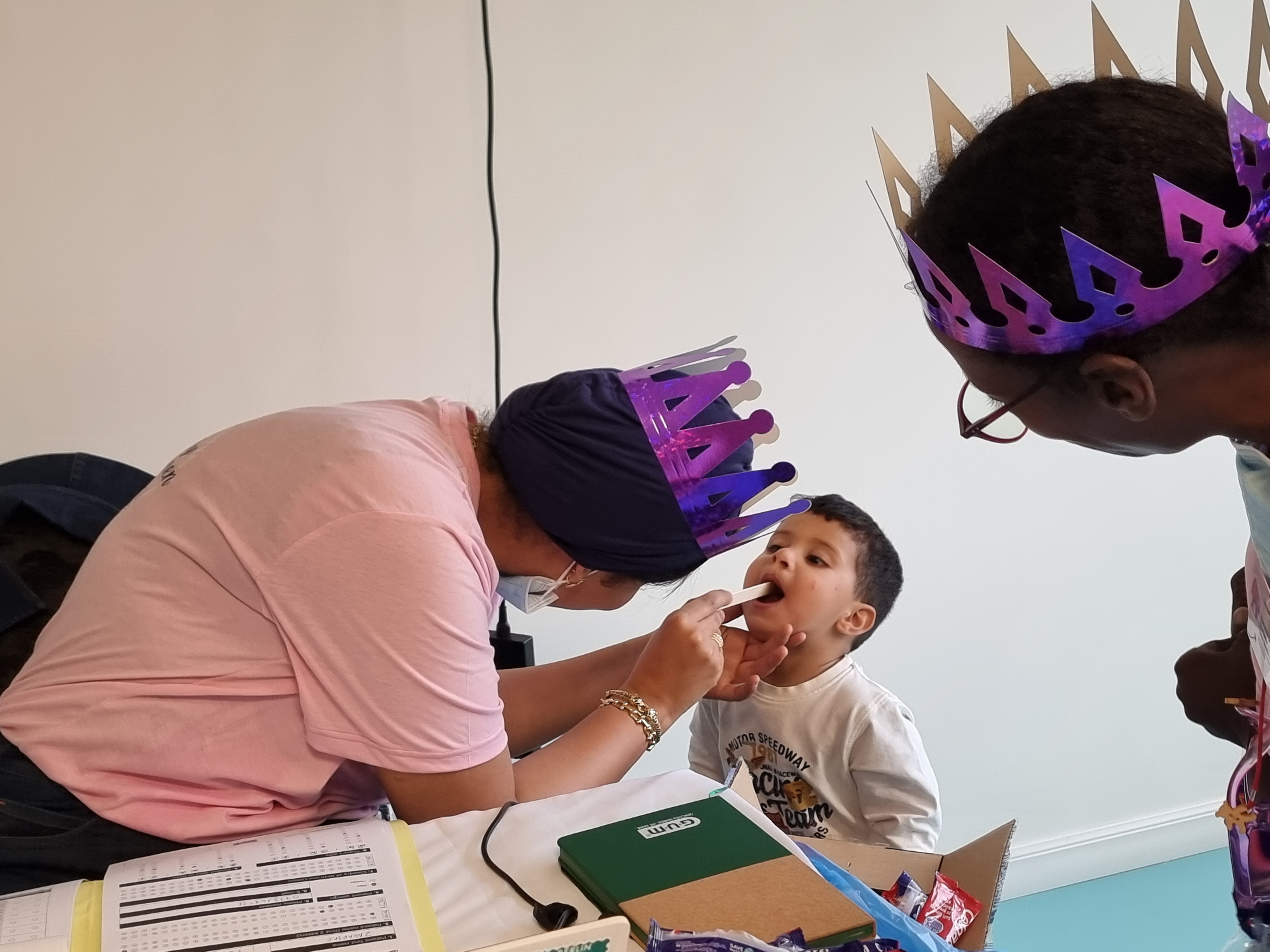 En vuxen med en lila krona undersöker ett barns mun medan en annan vuxen med krona tittar på. Det finns papper och en grön låda på bordet.