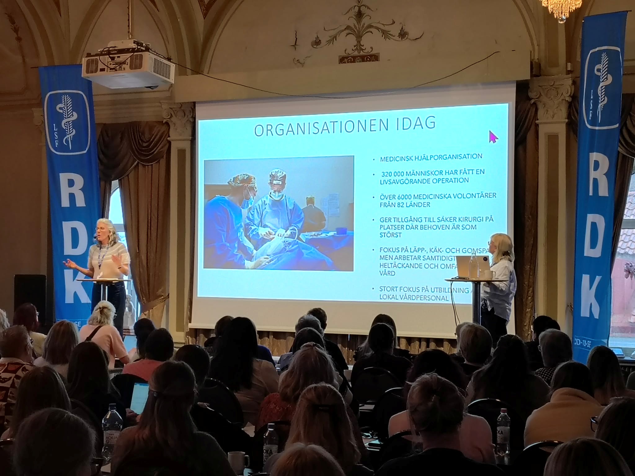 Två presentatörer står på en scen framför en publik, med en presentation om aktuell medicinsk organisation på en stor skärm.
