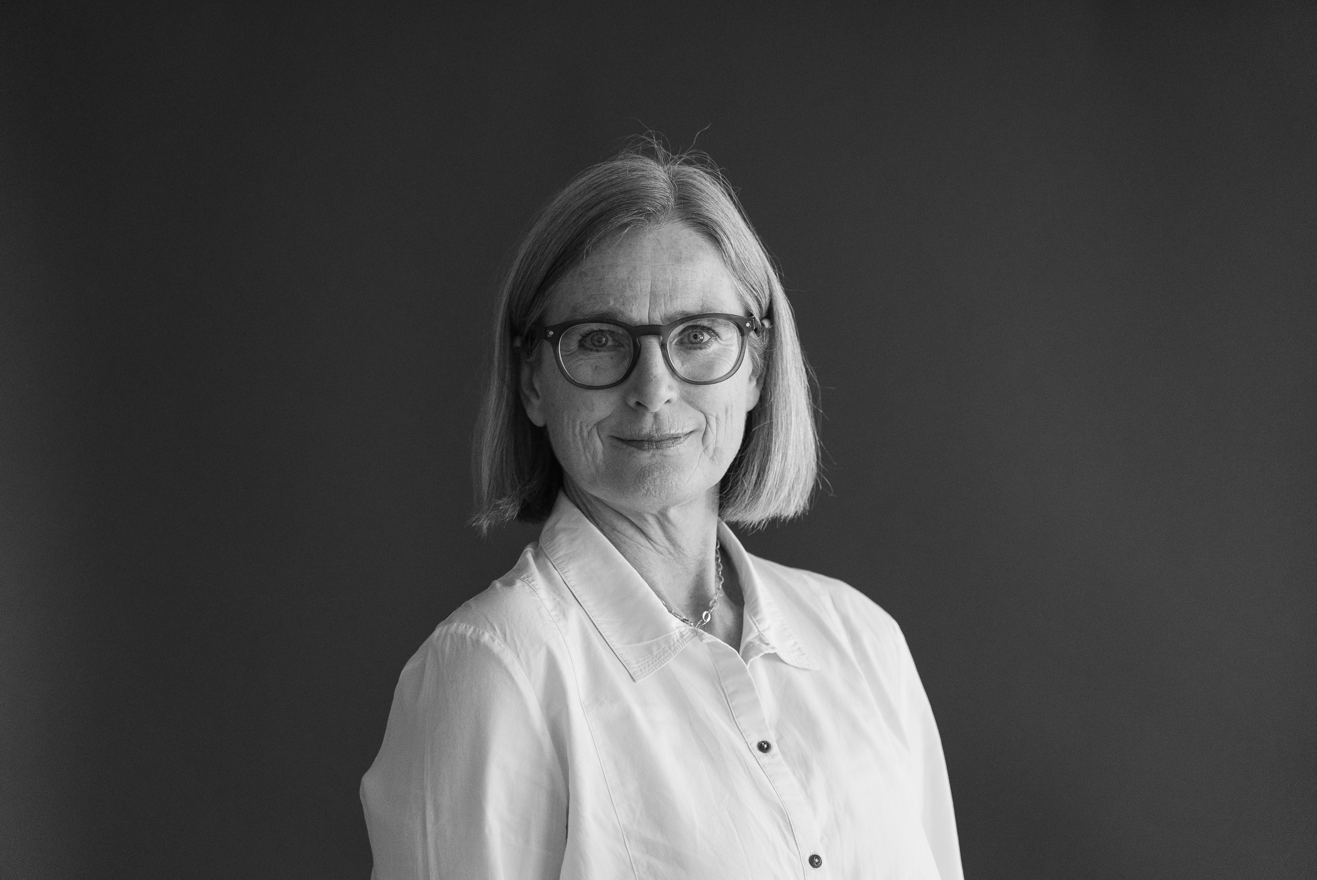 En kvinna med axellångt hår och glasögon, klädd i en skjorta med knappar, står framför en mörk bakgrund.