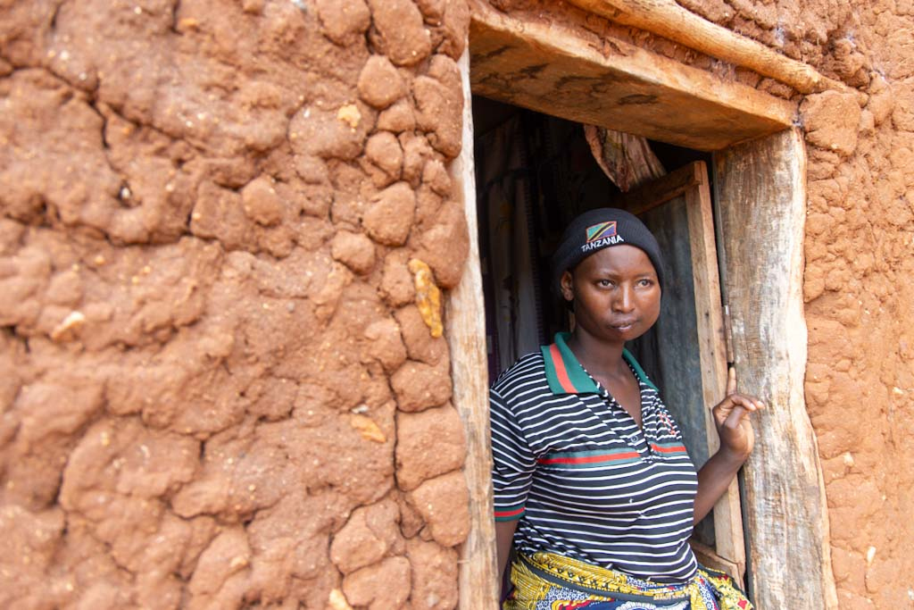 En person med randig skjorta och huvudscarf står i dörröppningen till en byggnad av lertegel och tittar ut.