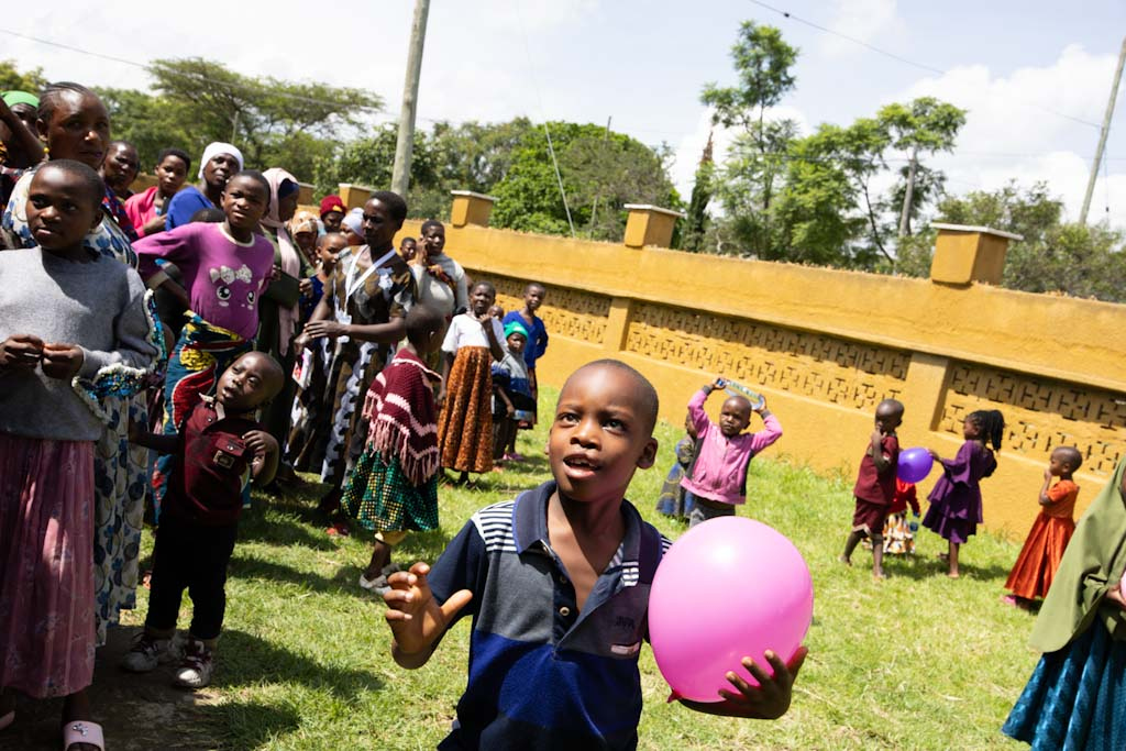 En pojke håller i en rosa ballong och står på gräset bland flera barn och vuxna nära en gul vägg.