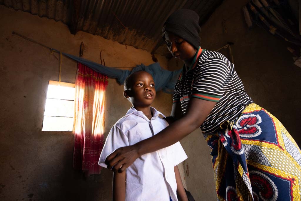 En kvinna hjälper en pojke att knäppa sin skjorta i ett rum med ett fönster och tyg hängande på väggen.
