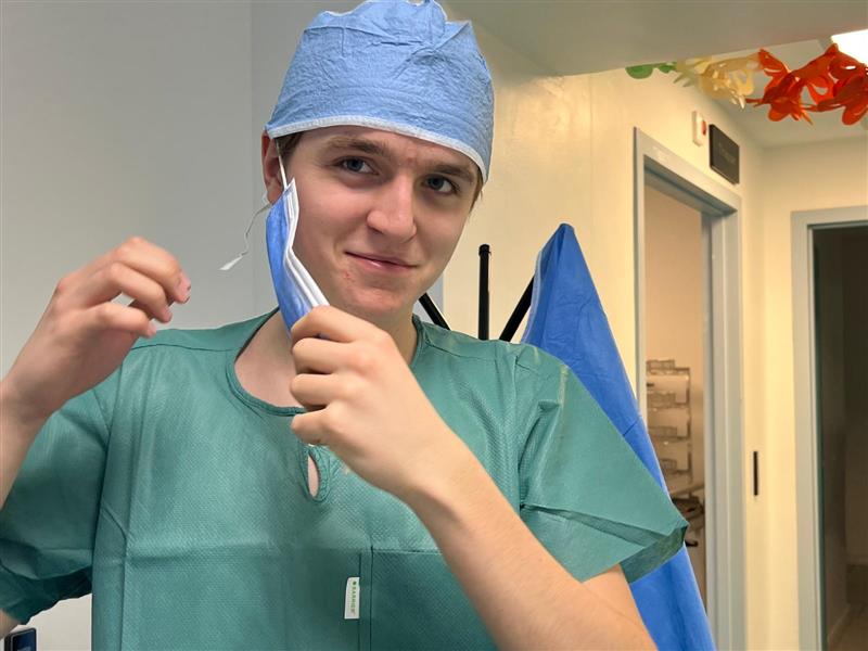 En person i gröna sjukhuskläder och blå operationsmössa tar på sig ett engångsmunskydd i en vårdmiljö.