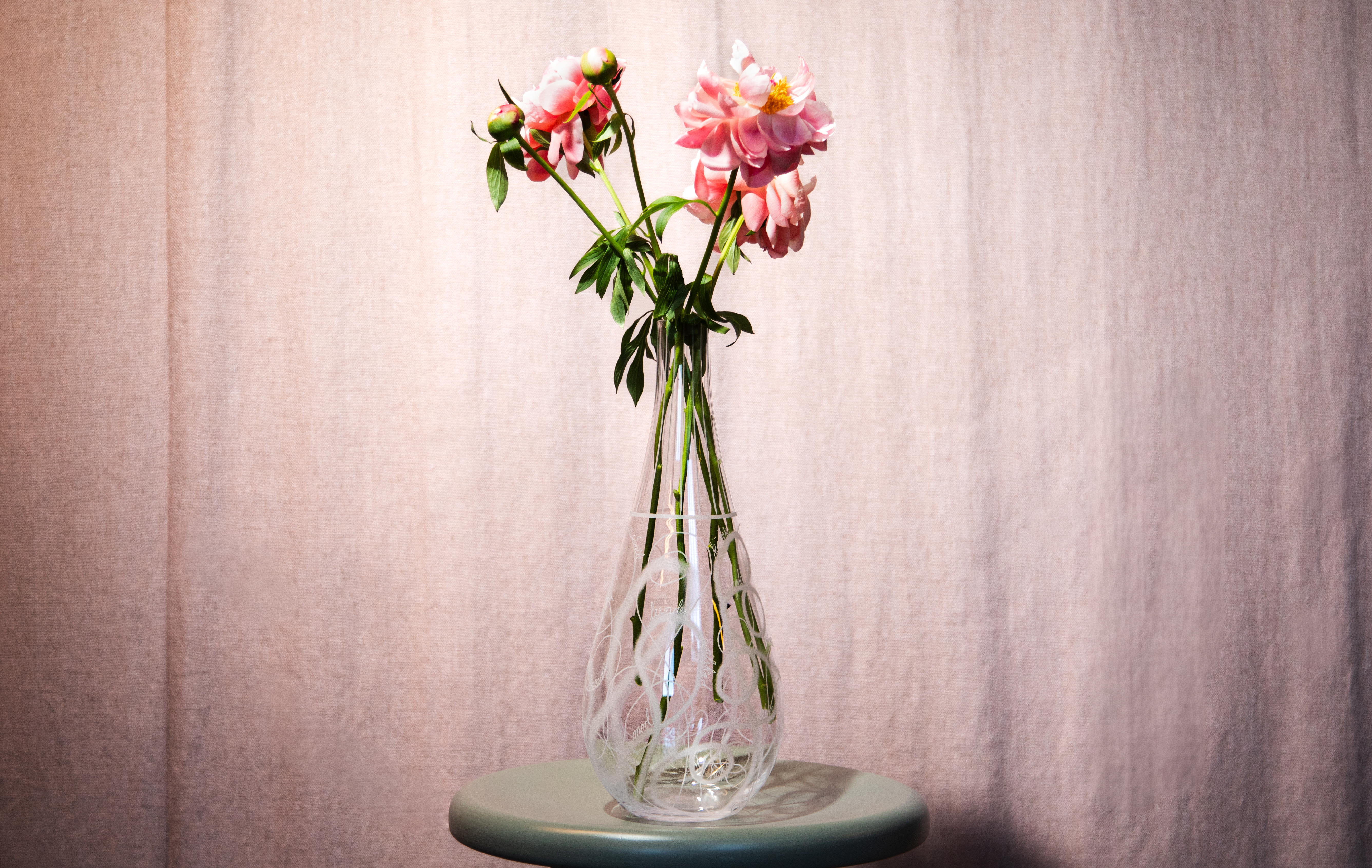 En glasvas med rosa blommor och gröna stjälkar står på ett runt grönt bord framför en ljusrosa gardin.