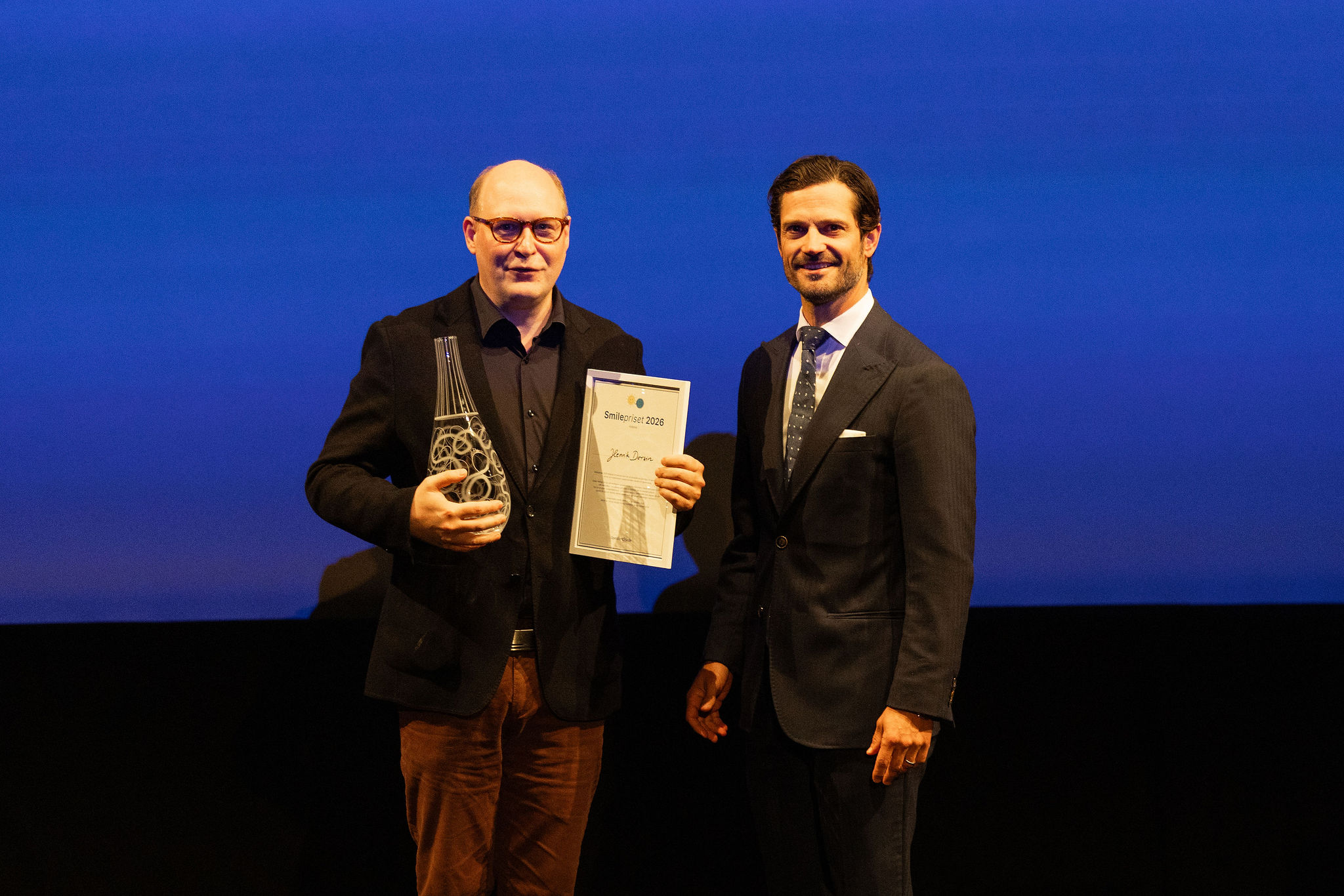 Henrik Dorsin och Prins Carl Philip står på en scen mot en blå bakgrund; den ene håller i ett glaspris och ett certifikat, den andre står bredvid honom, och båda vänder sig mot kameran.