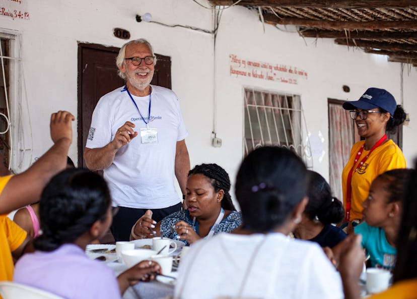 en mann med briller og hvit t-skjorte hvor det står «Operation Smile» prater til andre mennesker i hvite, oransje og lilla t-skjorter som sitter rundt et bord utenfor en hvitt murhus.