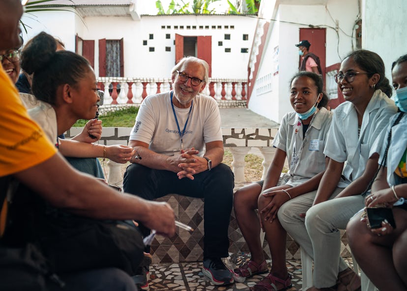 en mann med briller og hvit t-skjorte hvor det står «Operation Smile» ser på og lytter til en annen person som sitter i en sirkel med flere mennesker foran et hvitt murhus med rødmalte tredører.