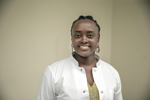 En person med vit labbrock och mönstrad skjorta står inomhus och ler mot kameran framför en ljus enfärgad bakgrund.