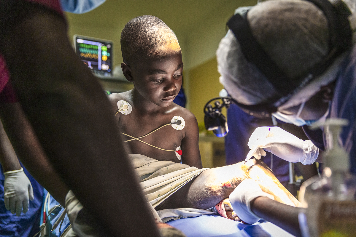 Ett barn ligger på en sjukhussäng med medicinska elektroder på bröstet medan vårdpersonal behandlar ett sår på benet.