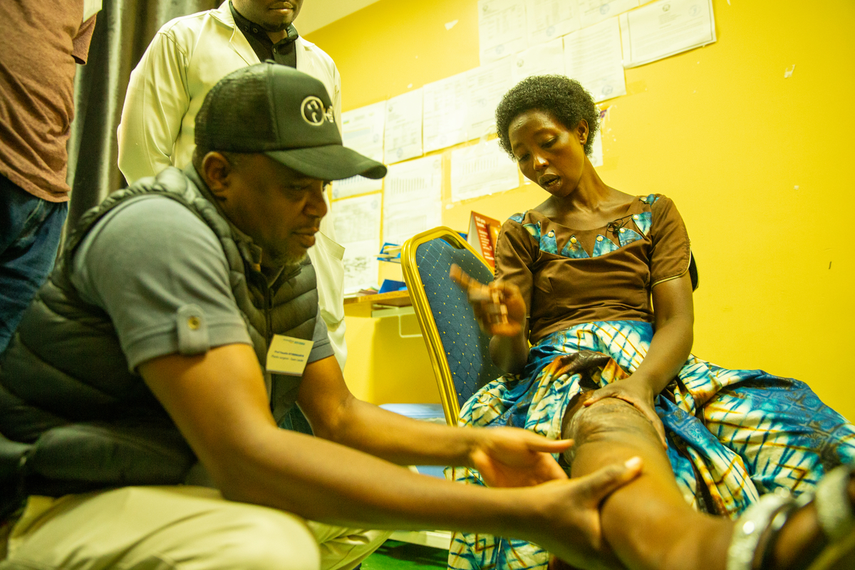 En sittande kvinna med bandagerat knä undersöks av en man med keps. En tredje person står bredvid i ett rum med gula väggar och papper på väggen.